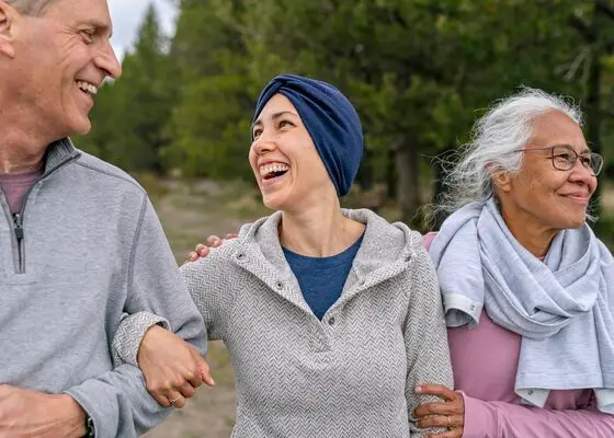 A young mixed race woman with cancer laughs while spending time outdoors with her supportive and loving parents. Their arms are linked and they are walking in a forested area. All of the individuals are smiling and feeling positive about the future.