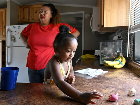 A young Black girl, wearing a yellow blouse, plays with seashells on a kitchen countertop. A Black woman, wearing a red blouse and red lipstick, stands with her hands on her hips in the background.